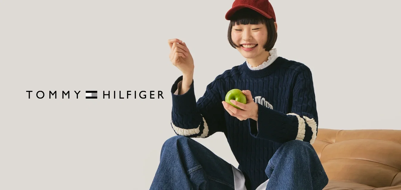 Junge Person mit roter Mütze, marineblauem Pullover und Jeans sitzt auf einem hellbraunen Ledersofa, hält einen grünen Apfel und lächelt vor einem schlichten Hintergrund.