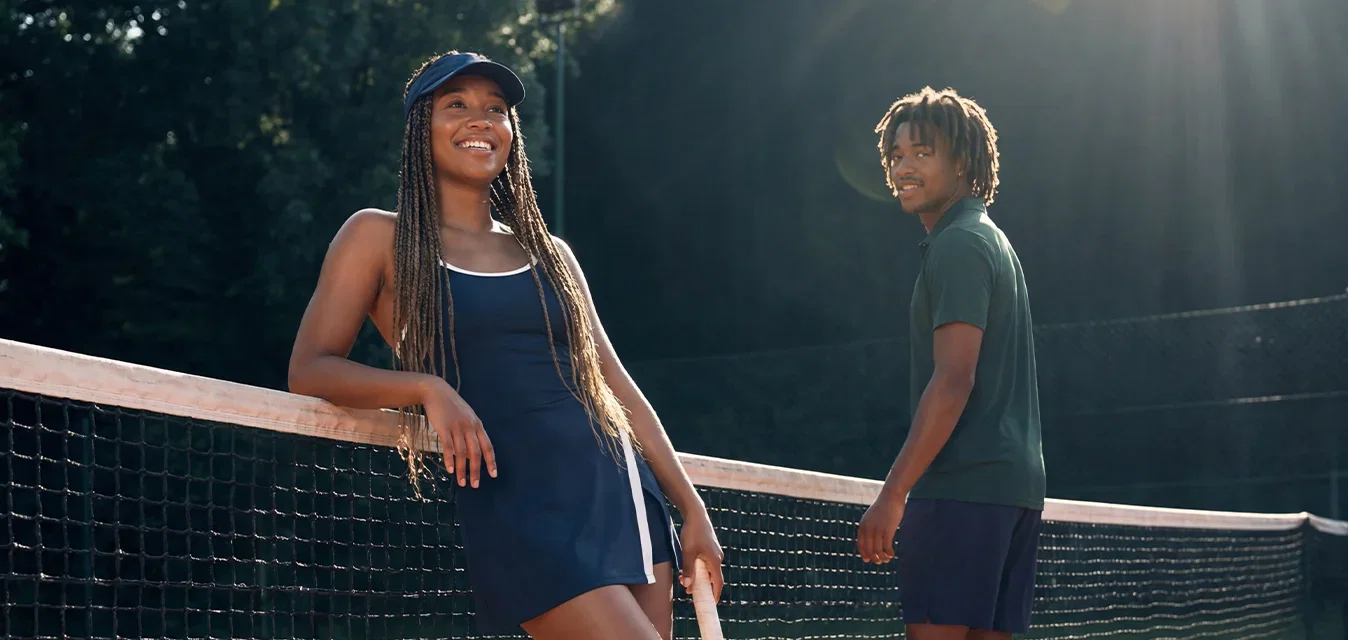 Zwei junge Personen stehen auf einem Sand-Tennisplatz am Netz, eine lehnt sich mit einem Schläger daran, im Hintergrund Sonnenlicht und Bäume.