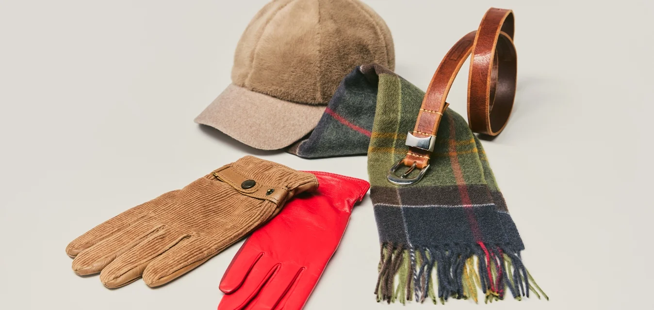 Braune Mütze, braune Cordhandschuhe, rote Lederhandschuhe, grün-blau kariertes Halstuch und brauner Ledergürtel auf hellem Hintergrund.
