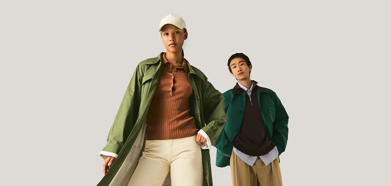 Zwei Models tragen geschichtete Herbstkleidung, darunter grüne Mäntel, braune Oberteile, beige und braune weite Hosen, vor einem einfarbigen hellen Hintergrund.