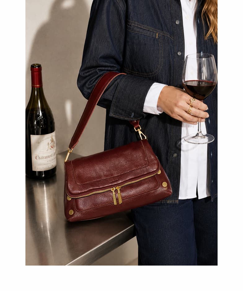 Model carrying a wine-toned pebble leather shoulder bag with gold hardware while holding a glass of red wine.