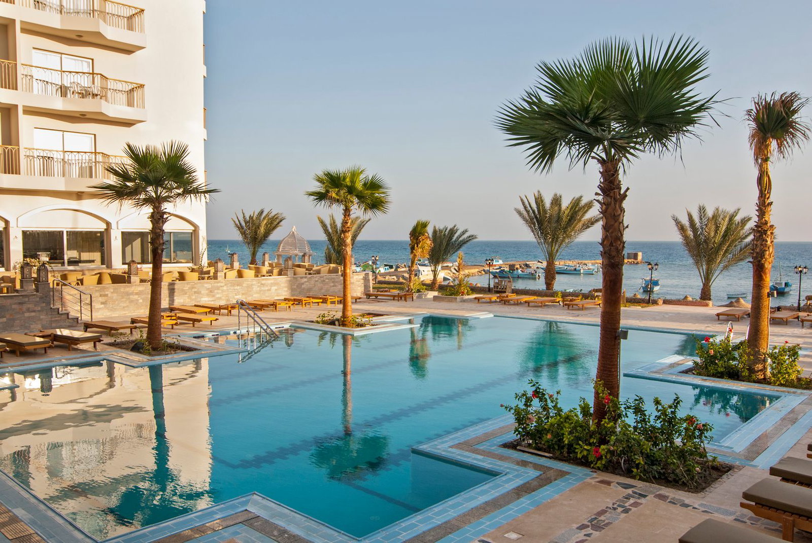 Seafront hotel swimming pool with palm trees, sun loungers and clear blue sea beyond, Mediterranean-style resort setting