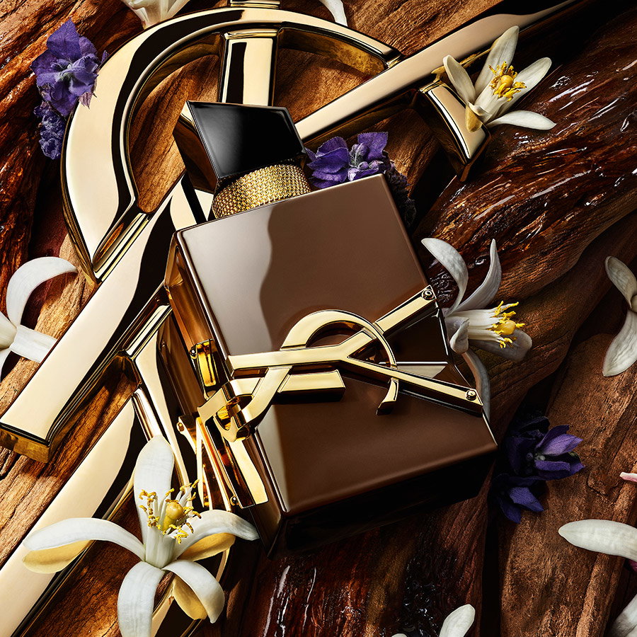 A brown YSL Libre fragrance bottle resting on wood alongside a golden logo, white orange blossoms, and purple lavender.