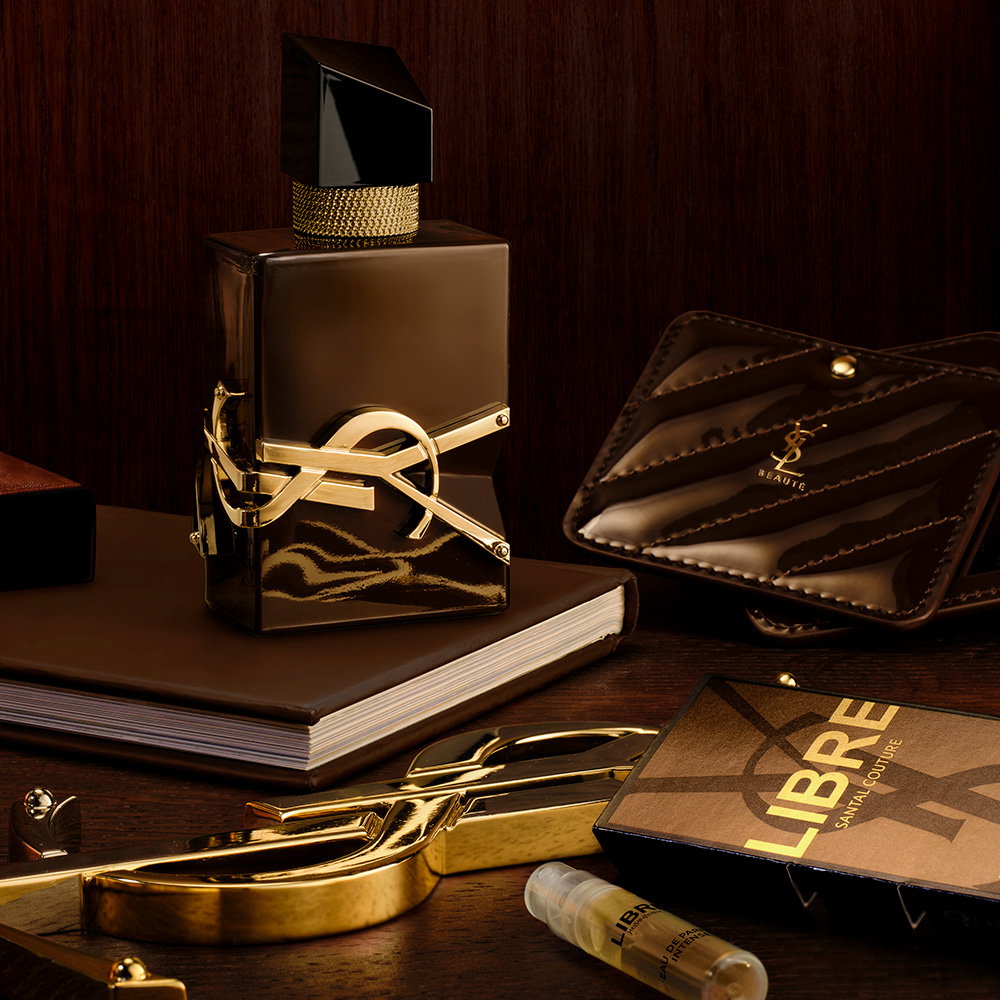A bottle of LIBRE SANTAL COUTURE perfume displayed on a dark wooden table next to its sample vial, a brown quilted leather pocket mirror, and golden YSL accessories.
