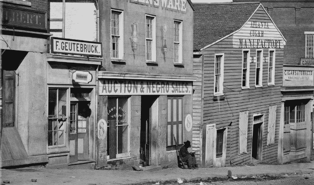 A 19th-century street scene featuring shops including an auction house with "Auction & Negro Sales" on its signage, and other nearby establishments.