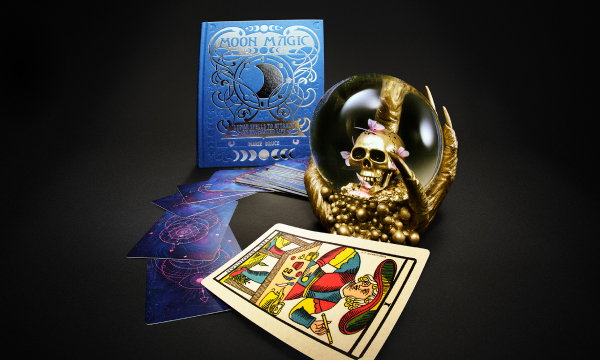 Crystal ball with a skull inside, tarot cards and a moon-themed book on a dark tabletop.