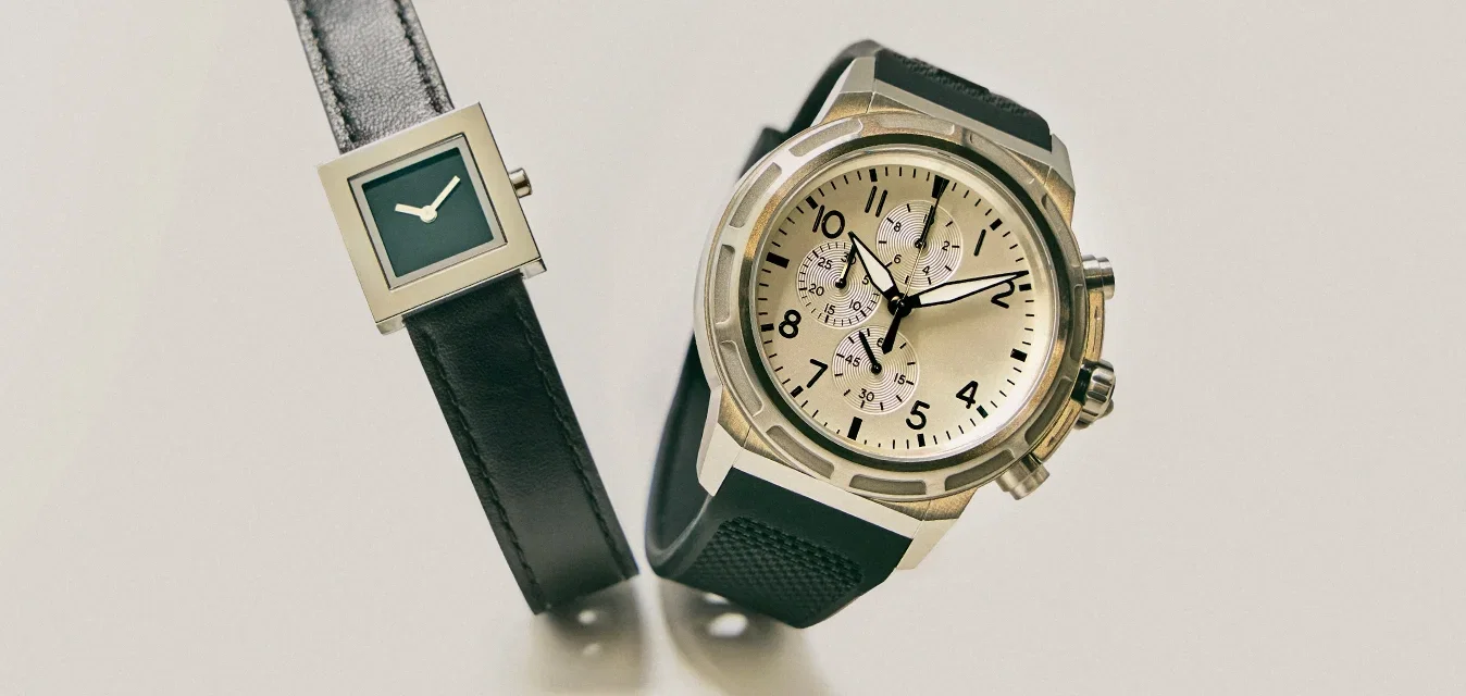 Zwei Armbanduhren mit schwarzen Armbändern auf einer hellen Oberfläche; eine hat ein quadratisches Zifferblatt, die andere ein rundes Chronographen-Zifferblatt mit Druckknöpfen.