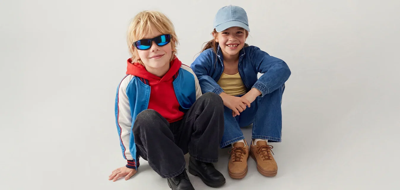 Zwei Kinder sitzen auf dem Boden vor einem hellen Hintergrund, eines trägt eine Sonnenbrille und einen roten Kapuzenpullover, das andere eine Mütze und eine Jeansjacke.