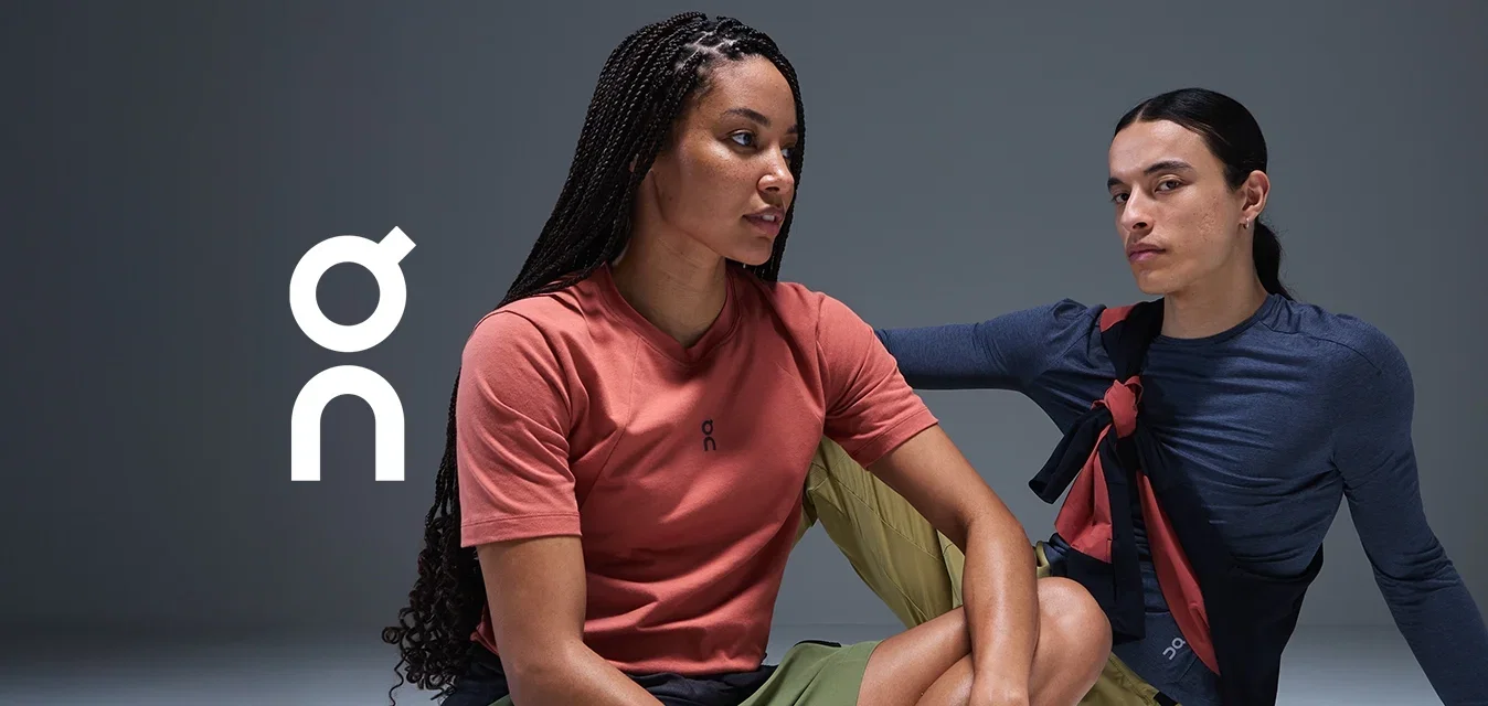 Zwei Personen sitzen vor einem hellen, glatten Hintergrund. Die eine trägt ein korallenfarbenes T-Shirt mit grünen Shorts, die andere ein marineblaues Langarmshirt mit ähnlichen Shorts.