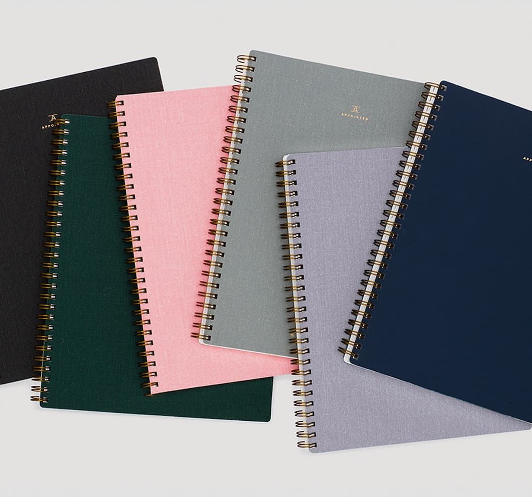 Signature Notebooks with brass wire-o binding in classic colorways are fanned out across a gray background.