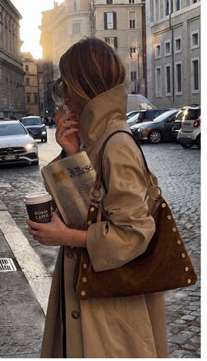 Model carrying a Kyle bag in Cinnamon Suede with gold rivets while walking on a city street.