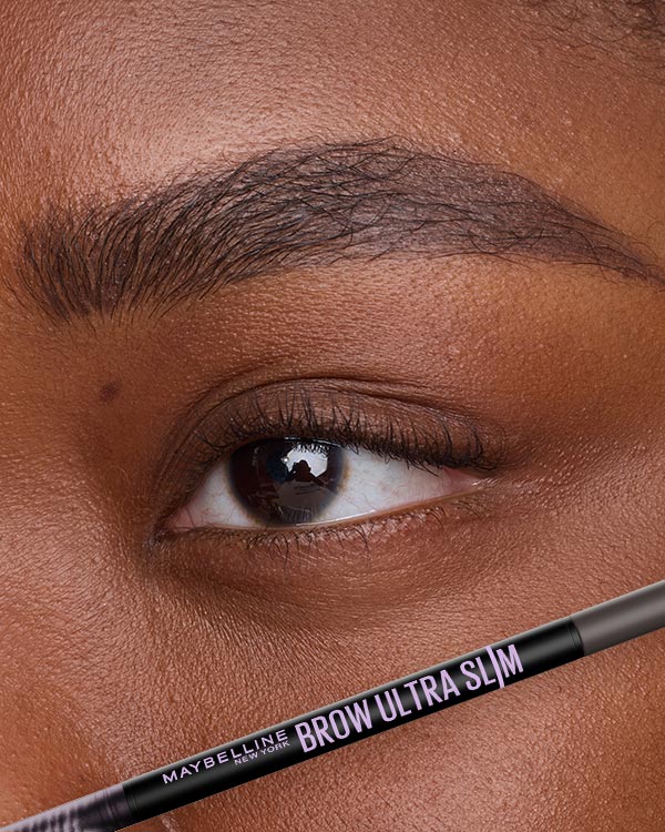 Close-up of a precisely filled dark brown eyebrow above a dark brown eye, with a Maybelline Brow Ultra Slim Pencil shown below to support a naturally shaped brow routine.