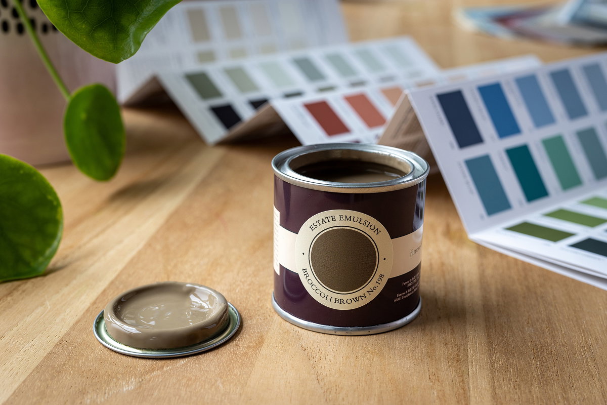Paint sample pot on a wooden table with colour swatches in the background, demonstrating how paint colours can be viewed and compared in natural light within a home setting.