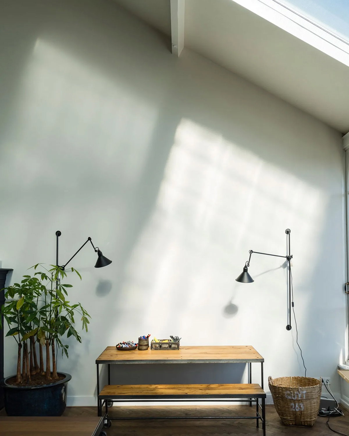 Wooden bench and table against a white wall, illuminated by soft sunlight from a high window, with potted plants and a woven basket on either side. 
