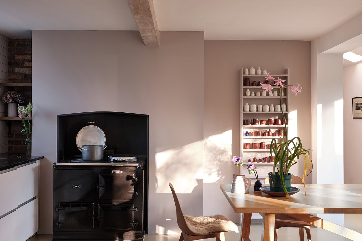 Sunlit dining area featuring a wooden table and chairs beside a black cast-iron stove, with shelves displaying paint samples and decorative items. 