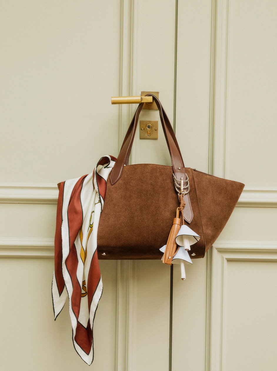 Braune Veloursleder-Tote-Bag von DOROTHEE SCHUMACHER mit Lederhenkeln, dekorativen Anhängern und gemustertem Seidentuch an einer cremefarbenen Tür.
