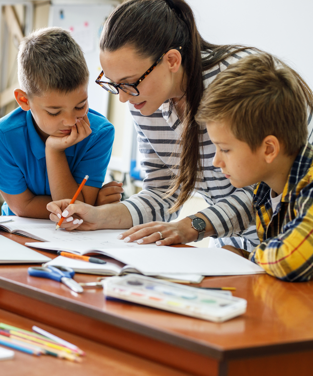 A teacher helps two young students with a classroom assignment, pointing to an open workbook with a pencil as the children look on.