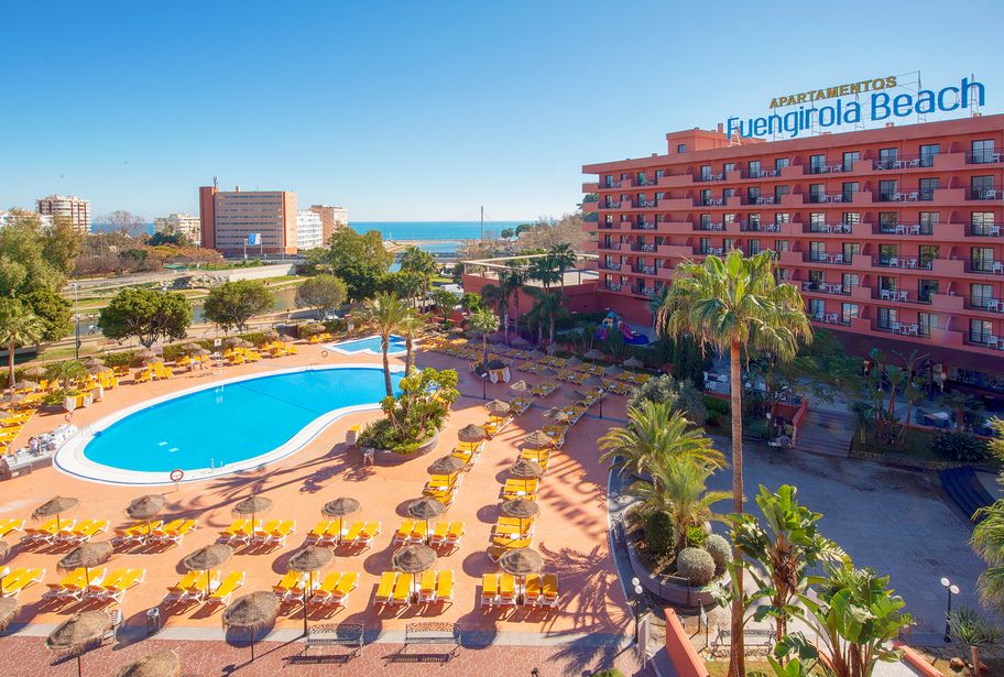Aerial view of Apartamentos Fuengirola Beach hotel with large outdoor swimming pool, rows of yellow sunloungers, palm trees and Mediterranean Sea beyond