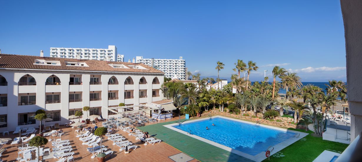 Seaside hotel resort with outdoor swimming pool, rows of sun loungers, palm trees and Mediterranean sea under a clear blue sky