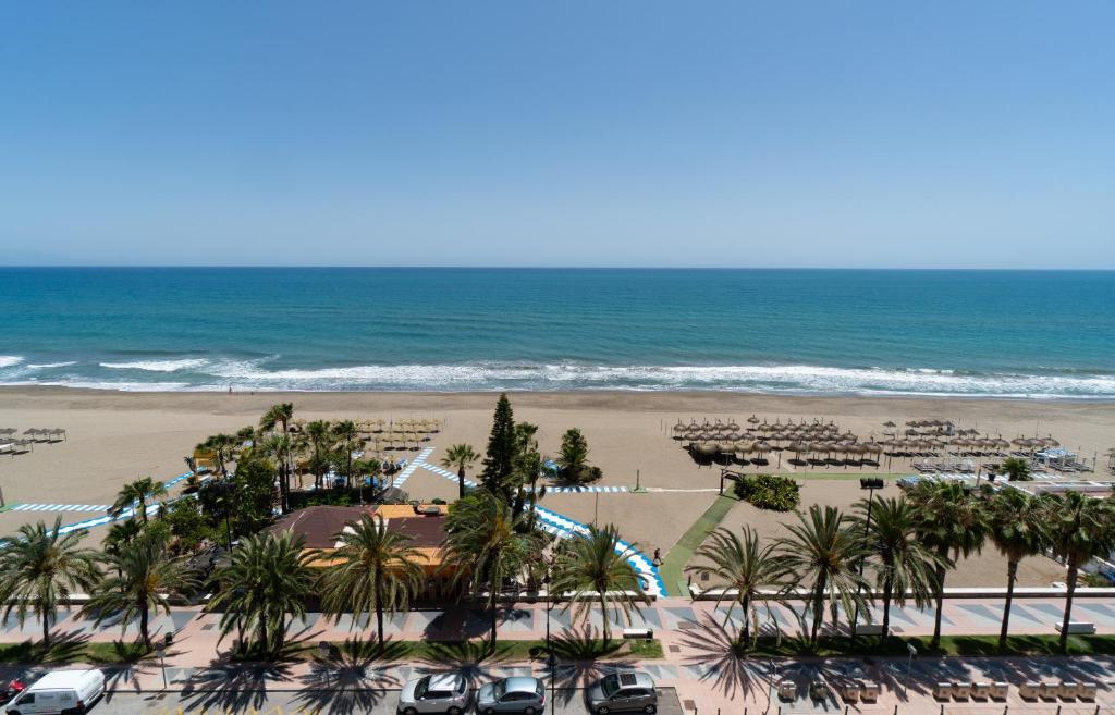 Aerial view of a sunny sandy beach with palm-lined promenade, rows of straw parasols, parked cars and blue sea