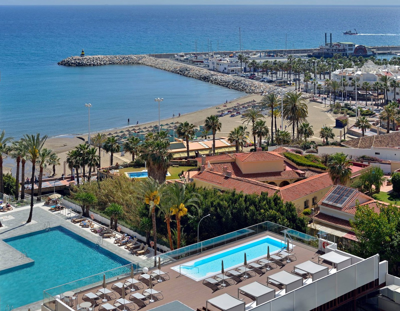 Aerial view of a Mediterranean beach resort with rooftop pools, sun loungers, palm trees, sandy beach, breakwater and blue sea