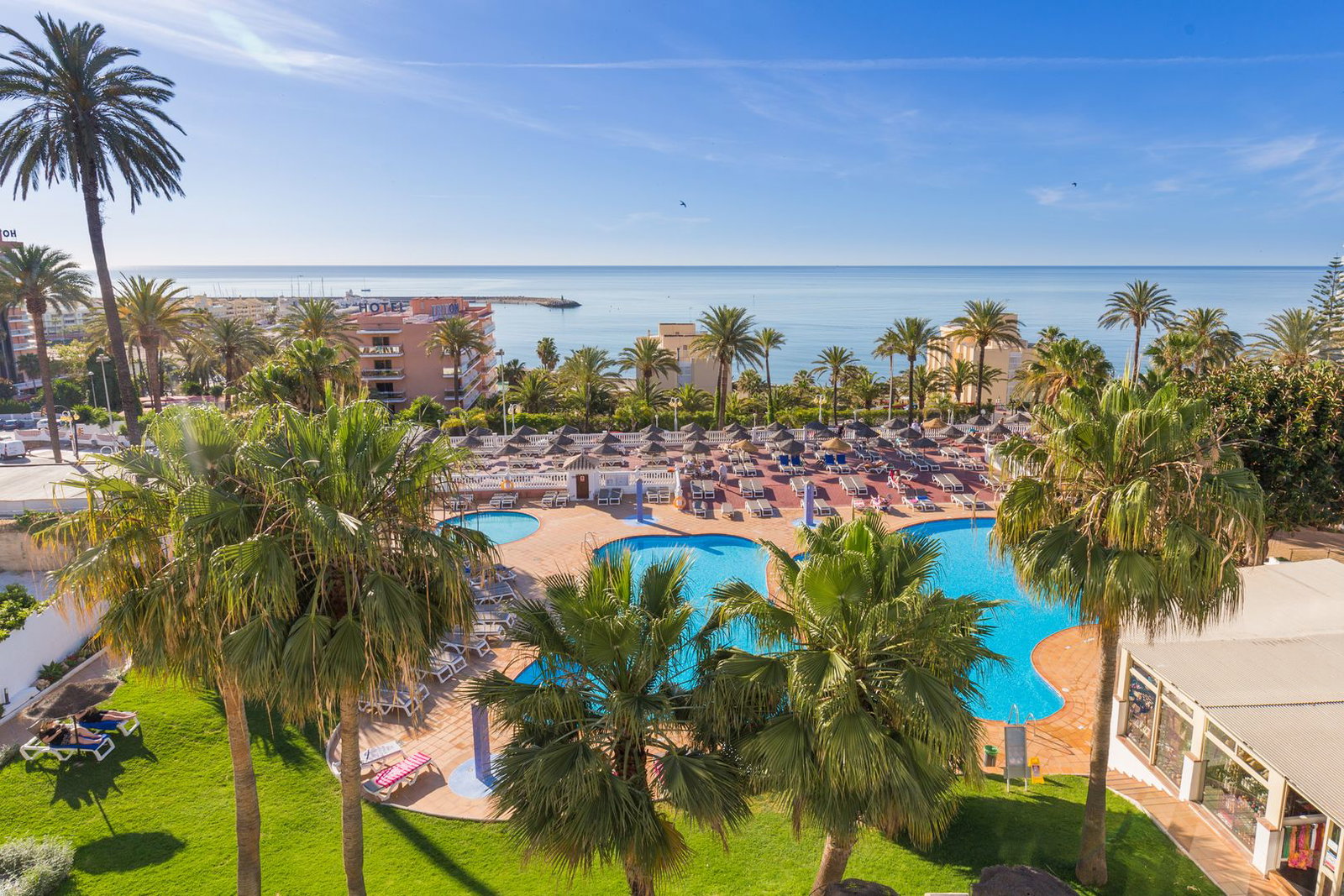 Resort swimming pool with palm trees and sun loungers overlooking the calm Mediterranean Sea under a clear blue sky