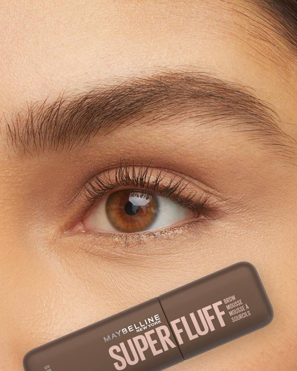 Close-up of a soft, full brown eyebrow above a light brown eye, with a Maybelline Superfluff Brow Mousse shown below to support a volumized brow routine.