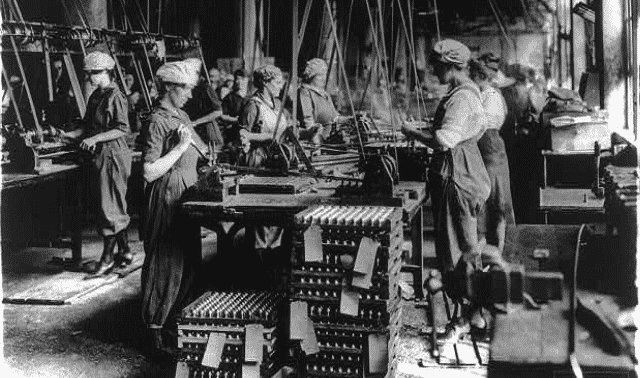 Workers in a factory assembling equipment, with various machinery and stacked parts around them. They are wearing uniforms and caps, indicative of early 20th-century industrial labor conditions.