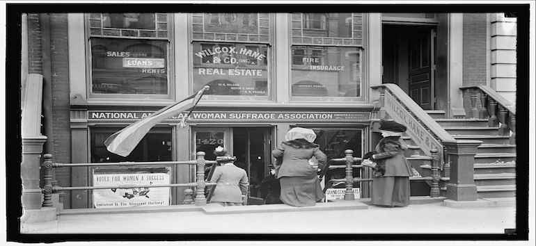 Three women are gathered outside the entrance of the National American Woman Suffrage Association office, with signs and flags advocating for women's right to vote.