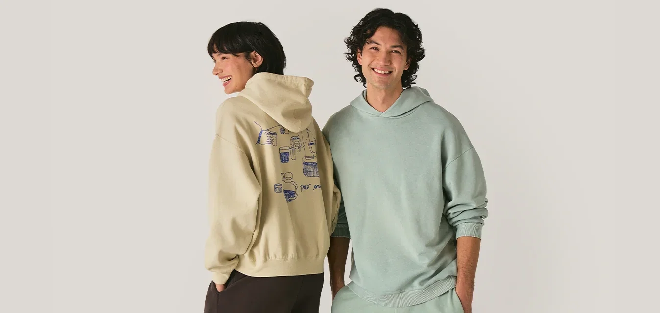 Lächelnder Mann und Frau in pastellfarbenen Hoodies und bequemen Hosen, die nebeneinander vor einem schlichten hellen Hintergrund stehen.