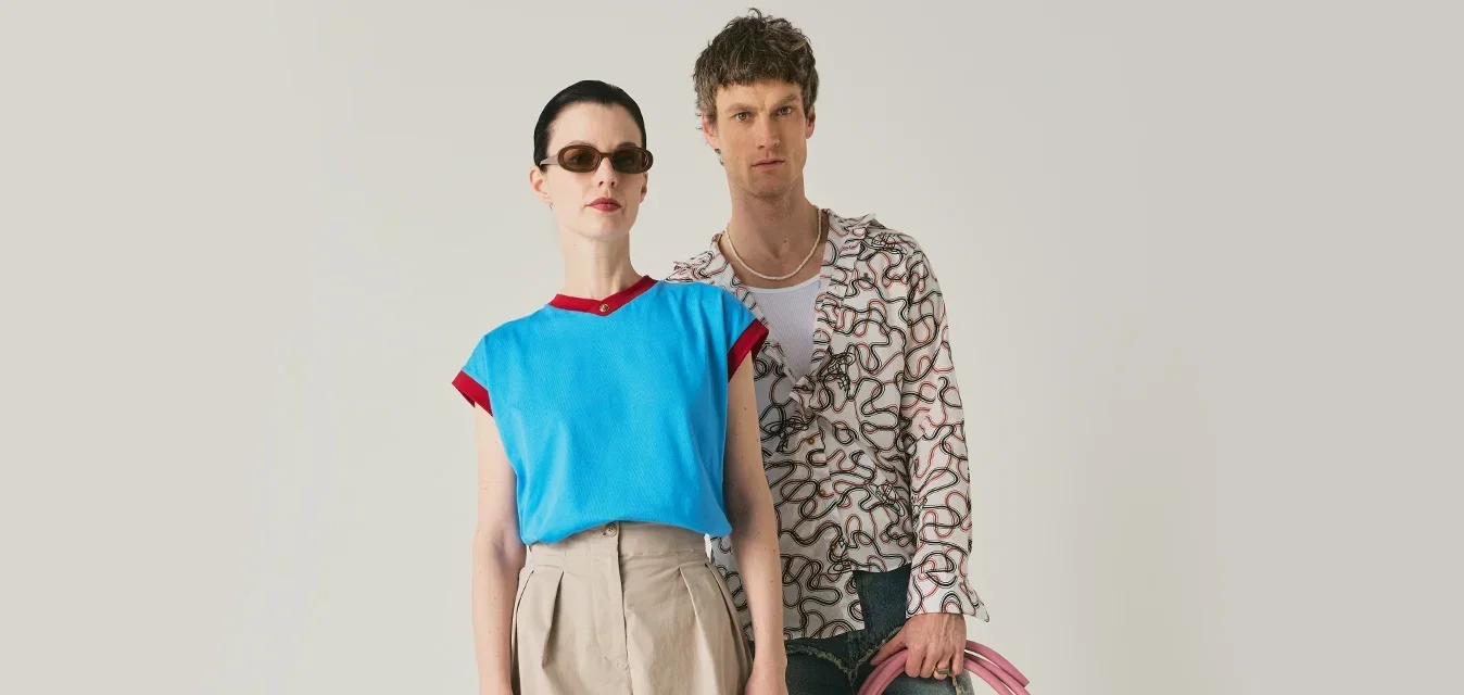 Zwei Models stehen mit rosa gewickelten Schläuchen; die Frau trägt ein blaues Top, beige Culottes, Sonnenbrille und hält eine gelbe, mit Nieten besetzte Tasche; der Mann trägt ein gemustertes Hemd und Jeansshorts.