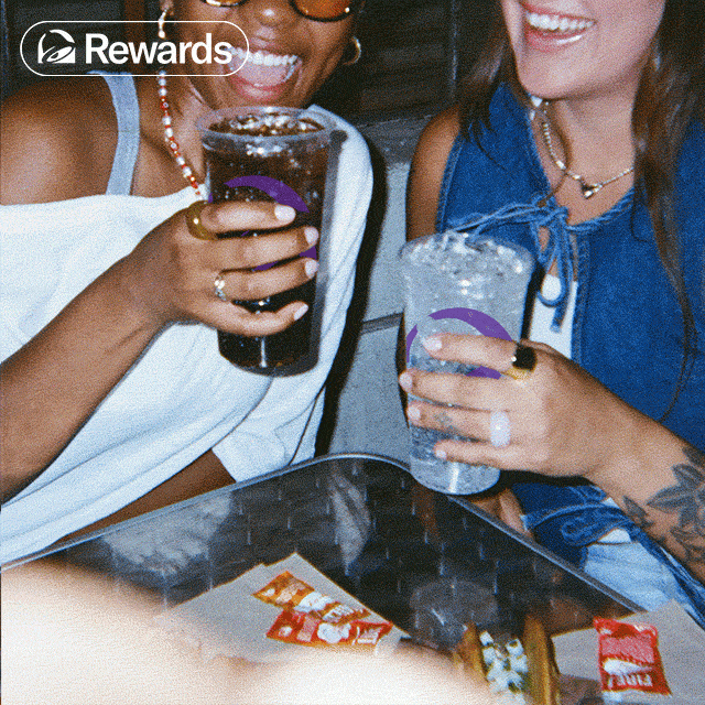 Two people sitting around a table, holding two beverages, and enjoying Taco Bell menu items.
