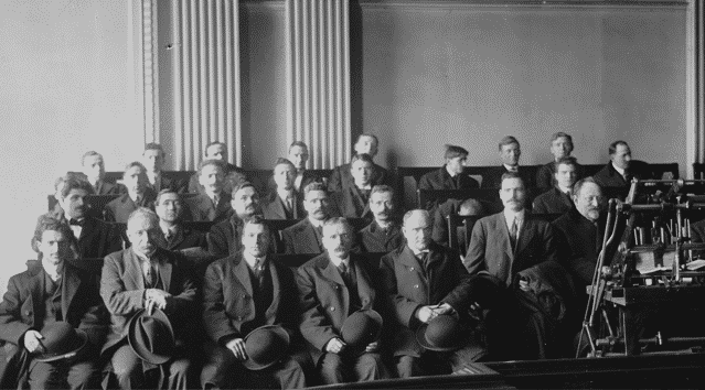 A group of men in early 20th-century attire sit and stand in a formal setting, some holding hats, with a machine on the right side of the image. Rows of columns are seen in the background.