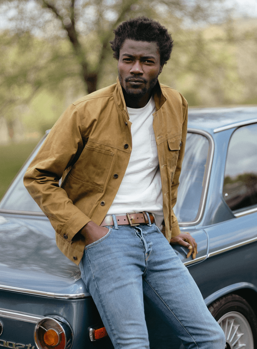 Man in a cream turtleneck sweater and blue jeans with a brown leather belt, standing on a sandy beach.