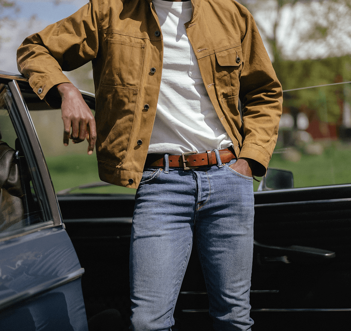 Close-up of a man wearing a brown leather belt with a brass buckle, paired with a black tee and green corduroy pants, hand in pocket.