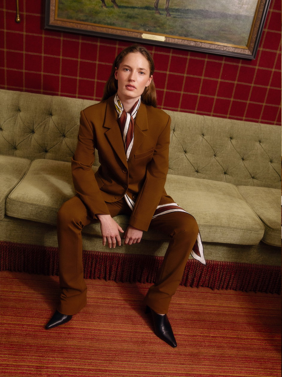 Model in einem braunen Hosenanzug von DOROTHEE SCHUMACHER mit gemustertem Seidentuch und schwarzen Stiefeletten, sitzend auf einem Vintage-Sofa.
