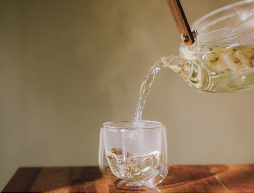 Tea being poured into a clear glass cup on a wooden table.