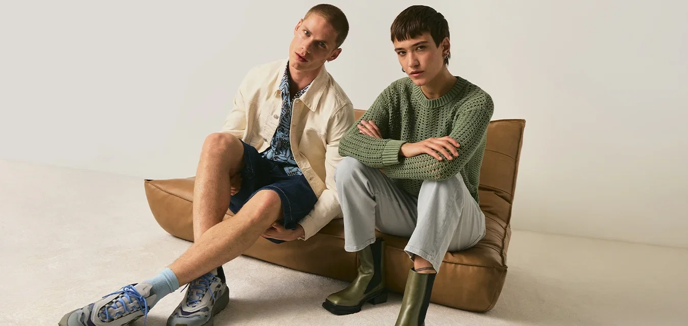 Zwei junge Erwachsene sitzen auf einem braunen Ledersofa auf dem Boden; einer trägt eine cremefarbene Jacke und Shorts, der andere einen grünen Pullover mit hellen Hosen und Stiefeln.
