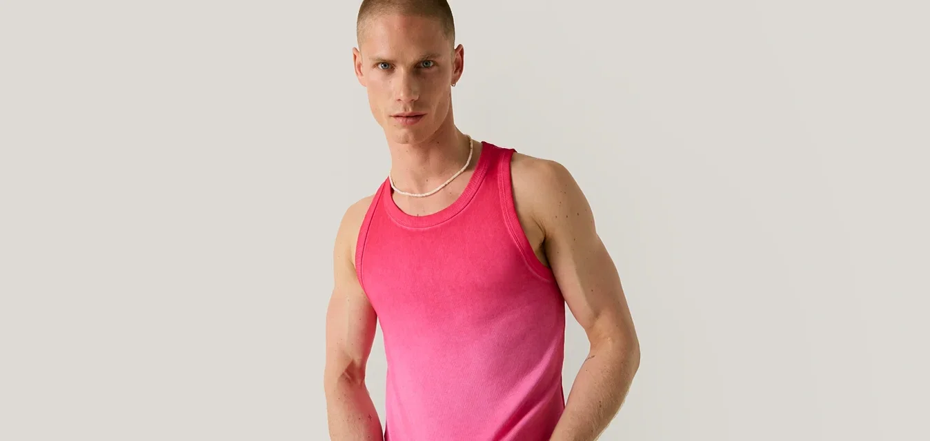 Mann mit kurzen Haaren, der ein ärmelloses pinkes Verlaufstanktop und beige Hosen trägt, steht vor einem einfarbigen hellgrauen Hintergrund.