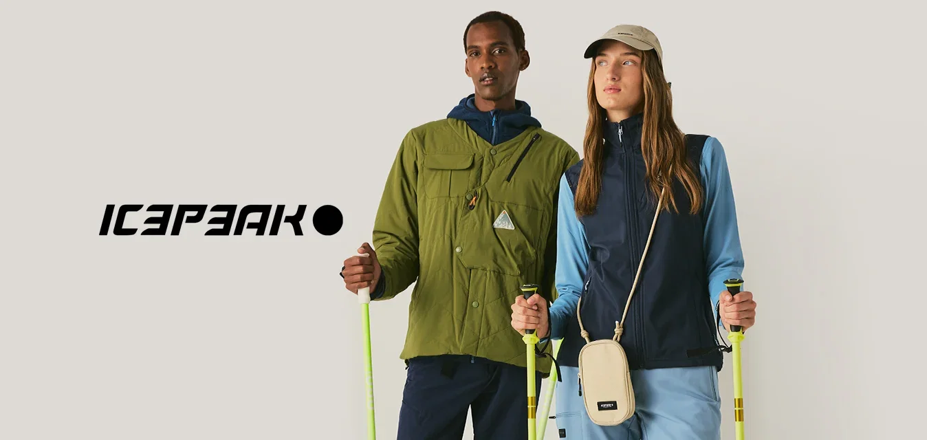 Zwei Personen mit Outdoor-Bekleidung und Wanderstöcken stehen vor einem einfarbigen Hintergrund und schauen nach vorne.