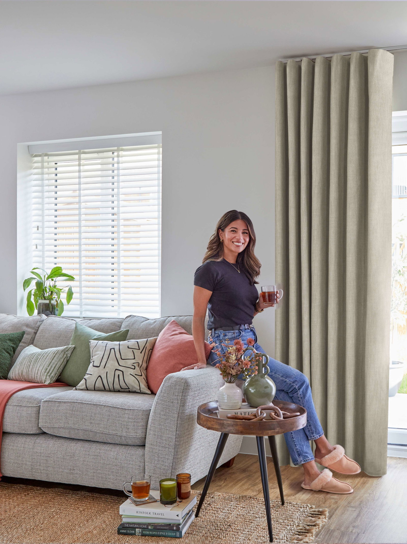 Person sitting on a sofa beside white blinds and full-length beige curtains framing French doors in a bright living room.