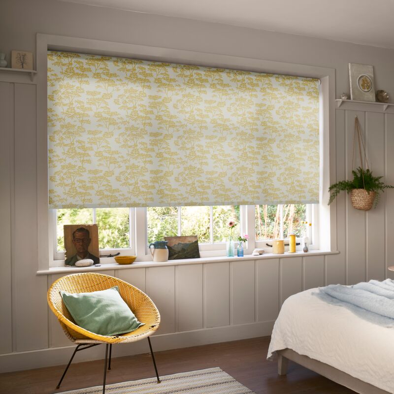 Yellow floral Roller blind filtering daylight at a bedroom window, complemented by soft neutral décor, a cushioned bed and woven chair.