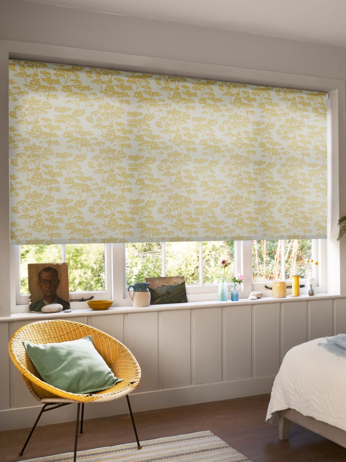 Yellow floral Roller blind filtering daylight at a bedroom window, complemented by soft neutral décor, a cushioned bed and woven chair.