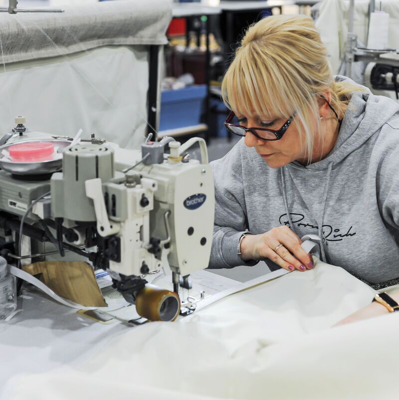 Person operating an industrial sewing machine, carefully guiding fabric through equipment in a busy textile workshop