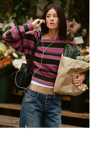 Young woman wearing pink and black striped crop sweater holding paper bag with earbuds around neck