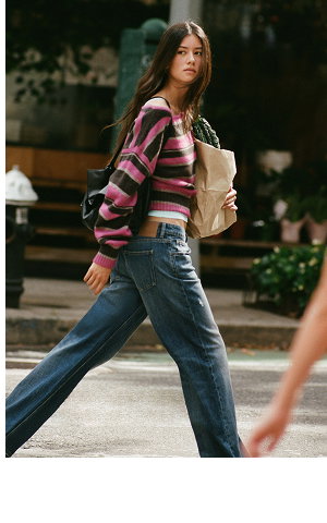 Young woman wearing a pink and black striped cropped cardigan, beige bucket bag, and distressed blue jeans