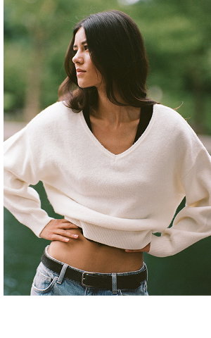 Woman wearing a cream-colored cropped v-neck sweater and blue jeans against a blurred green background