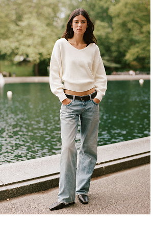 Woman wearing cream cropped sweatshirt, tan belt, and light blue jeans standing by a lake