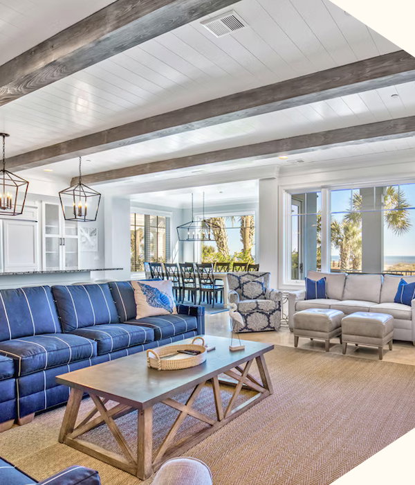 Spacious  seaside living area framed by vaulted beams, blue furnishings, sunlit windows, and coastal views beyond.