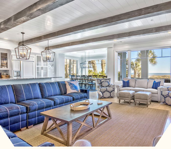 Spacious  seaside living area framed by vaulted beams, blue furnishings, sunlit windows, and coastal views beyond.
